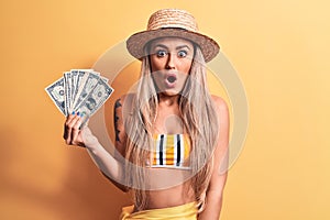 Young beautiful blonde woman wearing bikini and hat holding bunch of dollars banknotes scared and amazed with open mouth for