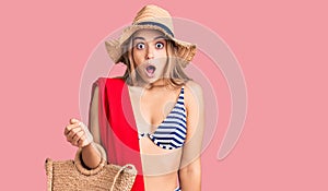 Young beautiful blonde woman wearing bikini and hat holding summer wicker handbag scared and amazed with open mouth for surprise,
