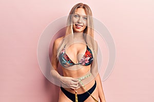 Young beautiful blonde woman wearing bikini using tape measure looking positive and happy standing and smiling with a confident