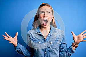 Young beautiful blonde woman wearing casual denim shirt and diadem over blue background crazy and mad shouting and yelling with