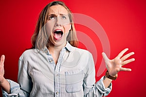 Young beautiful blonde woman wearing casual denim shirt over isolated red background crazy and mad shouting and yelling with
