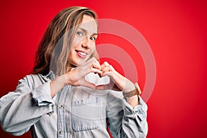 Young beautiful blonde woman wearing casual denim shirt over isolated red background smiling in love showing heart symbol and