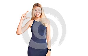 Young beautiful blonde woman wearing casual dress smiling and confident gesturing with hand doing small size sign with fingers