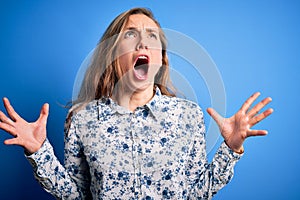 Young beautiful blonde woman wearing casual shirt standing over blue background crazy and mad shouting and yelling with aggressive