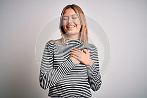 Young beautiful blonde woman wearing casual striped sweater over isolated background smiling with hands on chest with closed eyes