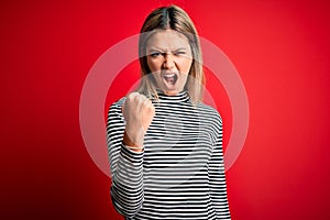Young beautiful blonde woman wearing casual striped sweater over red isolated background angry and mad raising fist frustrated and