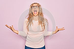 Young beautiful blonde woman wearing casual sweater and glasses over pink background crazy and mad shouting and yelling with