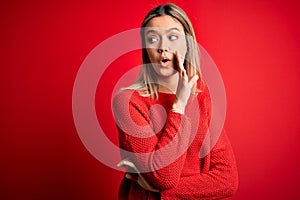 Young beautiful blonde woman wearing casual sweater over red isolated background hand on mouth telling secret rumor, whispering