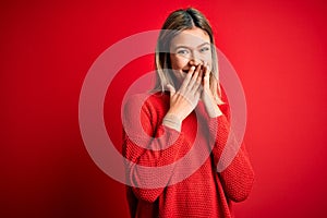 Young beautiful blonde woman wearing casual sweater over red isolated background laughing and embarrassed giggle covering mouth