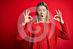 Young beautiful blonde woman wearing casual sweater over red isolated background looking surprised and shocked doing ok approval