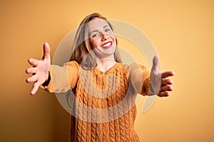 Young beautiful blonde woman wearing casual sweater standing over yellow background looking at the camera smiling with open arms