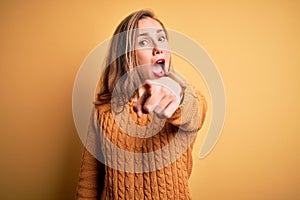 Young beautiful blonde woman wearing casual sweater standing over yellow background pointing displeased and frustrated to the