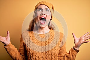 Young beautiful blonde woman wearing casual sweater and wool cap over yellow background crazy and mad shouting and yelling with