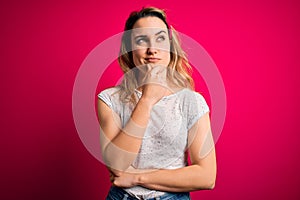 Young beautiful blonde woman wearing casual t-shirt standing over isolated pink background with hand on chin thinking about