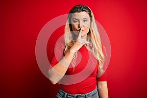 Young beautiful blonde woman wearing casual t-shirt standing over isolated red background asking to be quiet with finger on lips