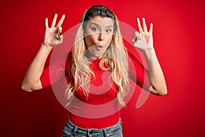 Young beautiful blonde woman wearing casual t-shirt standing over isolated red background looking surprised and shocked doing ok