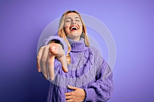 Young beautiful blonde woman wearing casual turtleneck sweater over purple background laughing at you, pointing finger to the