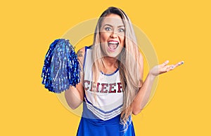 Young beautiful blonde woman wearing cheerleader uniform holding pompom celebrating victory with happy smile and winner expression