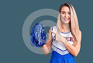Young beautiful blonde woman wearing cheerleader uniform holding pompom smiling happy pointing with hand and finger