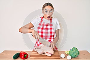 Young beautiful blonde woman wearing cook apron cutting chicken afraid and shocked with surprise and amazed expression, fear and