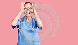 Young beautiful blonde woman wearing doctor uniform and stethoscope doing ok gesture like binoculars sticking tongue out, eyes