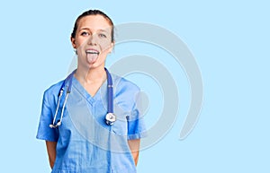 Young beautiful blonde woman wearing doctor uniform and stethoscope sticking tongue out happy with funny expression
