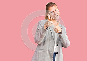Young beautiful blonde woman wearing elegant jacket pointing fingers to camera with happy and funny face