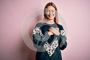 Young beautiful blonde woman wearing fashion sweater over pink  background smiling with hands on chest with closed eyes