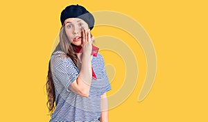 Young beautiful blonde woman wearing french beret and striped t-shirt hand on mouth telling secret rumor, whispering malicious