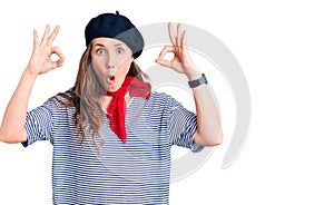 Young beautiful blonde woman wearing french beret and striped t-shirt looking surprised and shocked doing ok approval symbol with