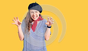 Young beautiful blonde woman wearing french beret and striped t-shirt smiling funny doing claw gesture as cat, aggressive and sexy