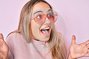 Young beautiful blonde woman wearing heart shaped sunglasses celebrating crazy and amazed for success with open eyes screaming