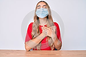 Young beautiful blonde woman wearing medical mask sitting on the table smiling with hands on chest, eyes closed with grateful