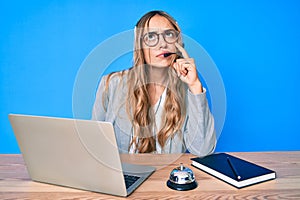 Young beautiful blonde woman wearing operator headset at the call center office serious face thinking about question with hand on