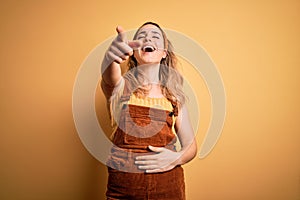 Young beautiful blonde woman wearing overalls and diadem standing over yellow background laughing at you, pointing finger to the