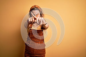 Young beautiful blonde woman wearing overalls and diadem standing over yellow background Rejection expression crossing fingers