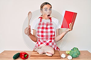 Young beautiful blonde woman wearing professional baker apron reading cooking recipe book afraid and shocked with surprise and