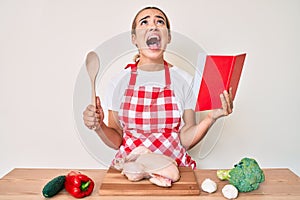 Young beautiful blonde woman wearing professional baker apron reading cooking recipe book angry and mad screaming frustrated and