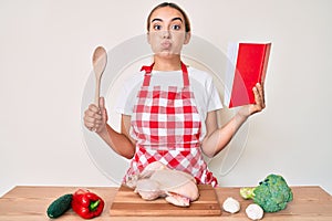 Young beautiful blonde woman wearing professional baker apron reading cooking recipe book puffing cheeks with funny face