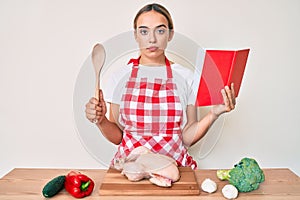 Young beautiful blonde woman wearing professional baker apron reading cooking recipe book relaxed with serious expression on face