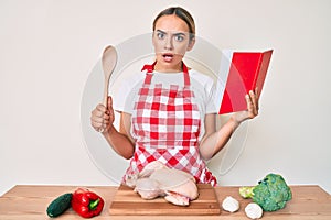 Young beautiful blonde woman wearing professional baker apron reading cooking recipe book in shock face, looking skeptical and