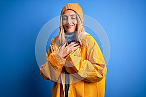 Young beautiful blonde woman wearing raincoat for rainy weather over blue background smiling with hands on chest with closed eyes