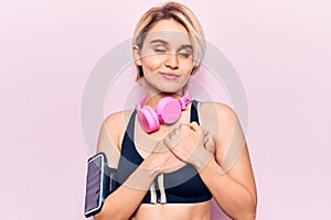 Young beautiful blonde woman wearing sportswear using headphones smiling with hands on chest with closed eyes and grateful gesture