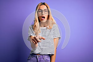 Young beautiful blonde woman wearing striped t-shirt and glasses over purple background pointing displeased and frustrated to the