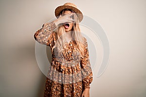 Young beautiful blonde woman wearing summer dress and hat over isolated white background peeking in shock covering face and eyes
