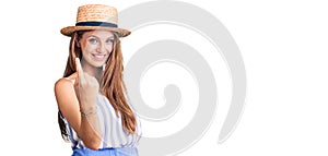 Young beautiful blonde woman wearing summer hat beckoning come here gesture with hand inviting welcoming happy and smiling