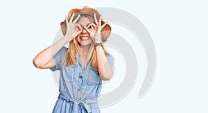 Young beautiful blonde woman wearing summer hat and dress doing ok gesture like binoculars sticking tongue out, eyes looking