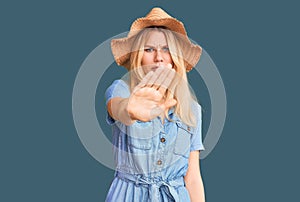 Young beautiful blonde woman wearing summer hat and dress doing stop sing with palm of the hand