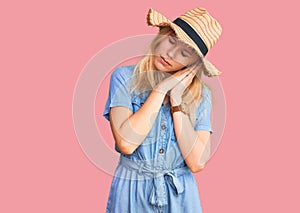 Young beautiful blonde woman wearing summer hat and dress sleeping tired dreaming and posing with hands together while smiling