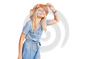 Young beautiful blonde woman wearing summer hat and dress very happy and smiling looking far away with hand over head